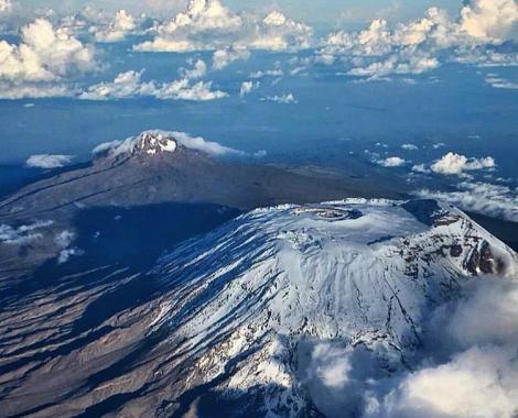 An aerial view looking down at the massive, snow-capped peak of Mount Kilimanjaro rising above a blanket of clouds.