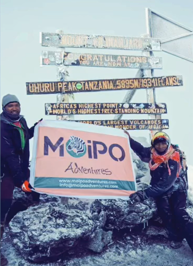 Two triumphant climbers celebrating at Uhuru Peak, holding up a branded Moipo banner at the summit of Mount Kilimanjaro. Instagram post